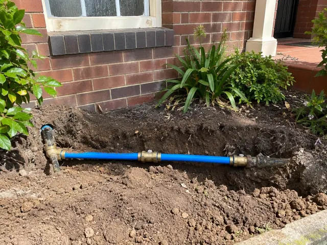 Open trench with a newly repaired blue water pipe beside a brick house wall in Gordon